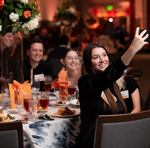 A woman holds out a cell phone to take a selfie of people seated at a table during the Alumni Awards Gala