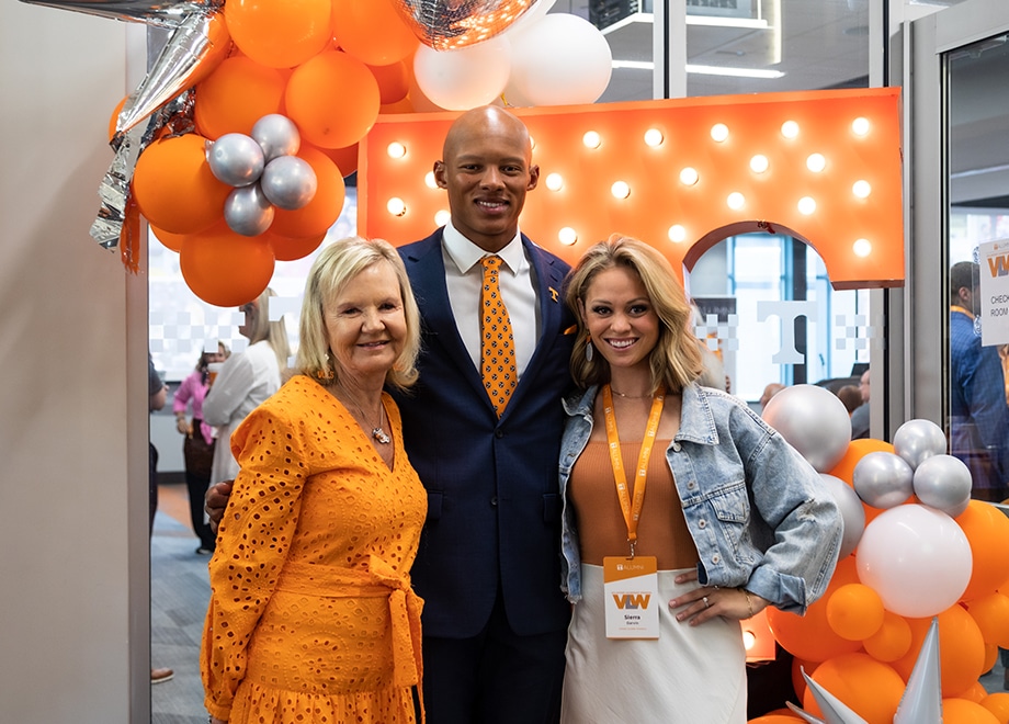 Two women and a man smile for a group photo in front of an orange T and orange-and-white balloons