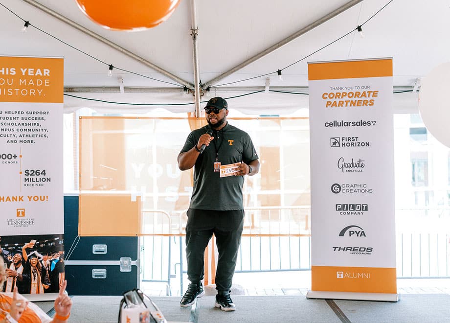 A speaker during a Tennessee Tailgate stands next to a sign recognizing Alumni Corporate Partners