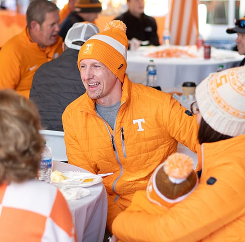 A man wearing a Tennessee Volunteers winter hat and orange jacket sits at a table and chats with people out of the frame
