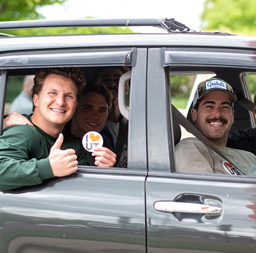 Two students in a vehicle smile for a photo during an I Heart UT week event. One student holds a button and gives a thumbs up.