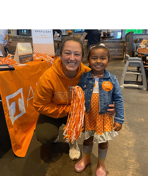 An adult and a child decked out in Tennessee Volunteer gear smile for a photo in front of a UT Knoxville alumni table