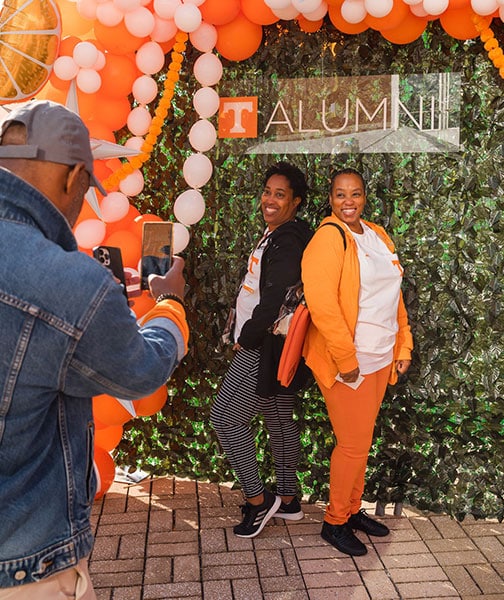 A man takes a photo of two women against a UT Knoxville Alumni photo backdrop during the Homecoming tailgate