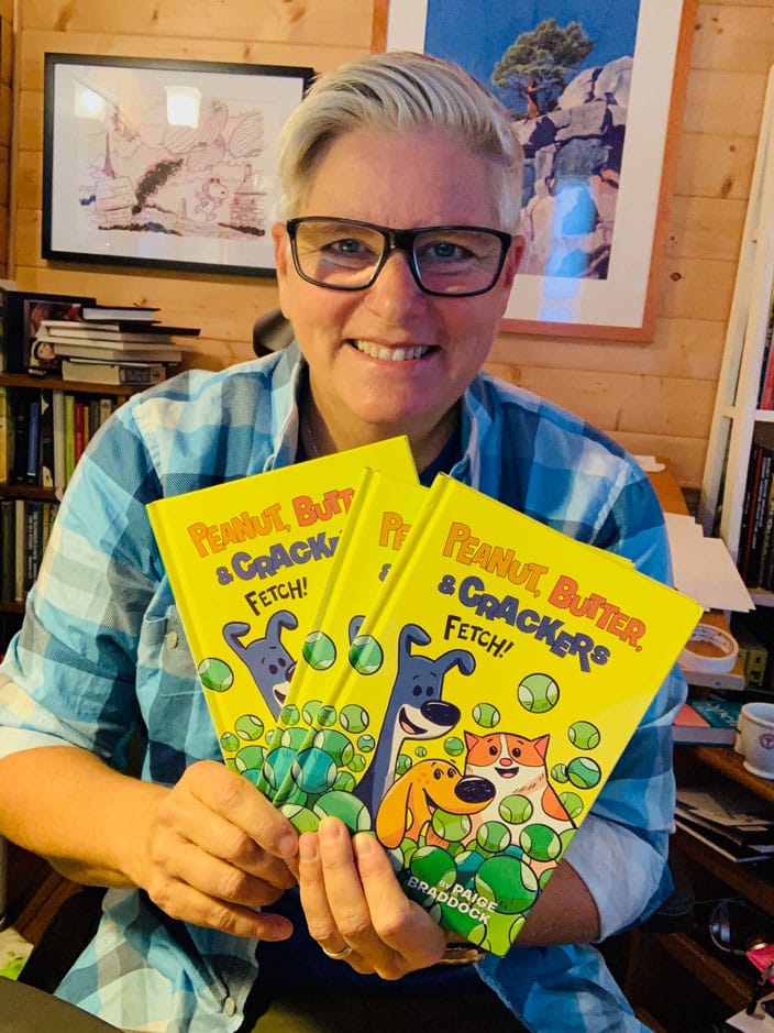 braddock-paige-peanut-butter-and-crackers-books Paige Braddock holds three copies of her Peanut, Butter, and Crackers Fetch book. The book is bright yellow with comic characters of two dogs and a cat.