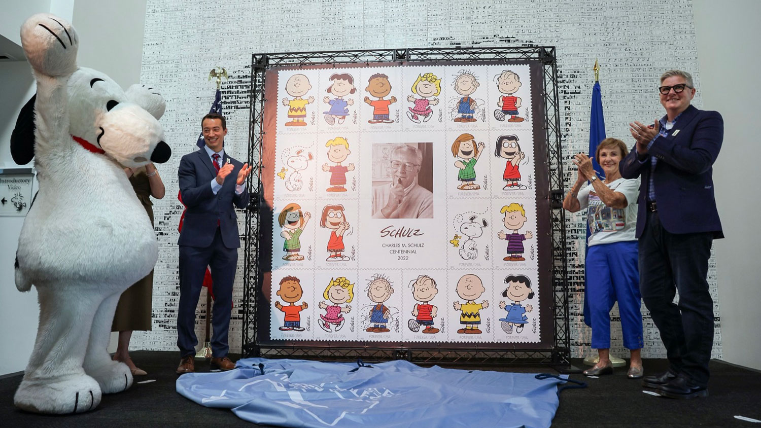 braddock-snoopy_stamp_event Snoopy stands to the left of the frame, and Paige Braddock stands to the right. In the center is a giant stamp honoring the Charles M Schulz Centennial 2022.