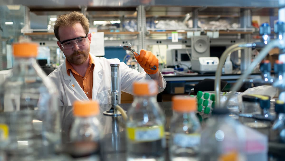 Tyler Myers, wearing safety goggles, orange gloves, and a lab coat, working in a lab.