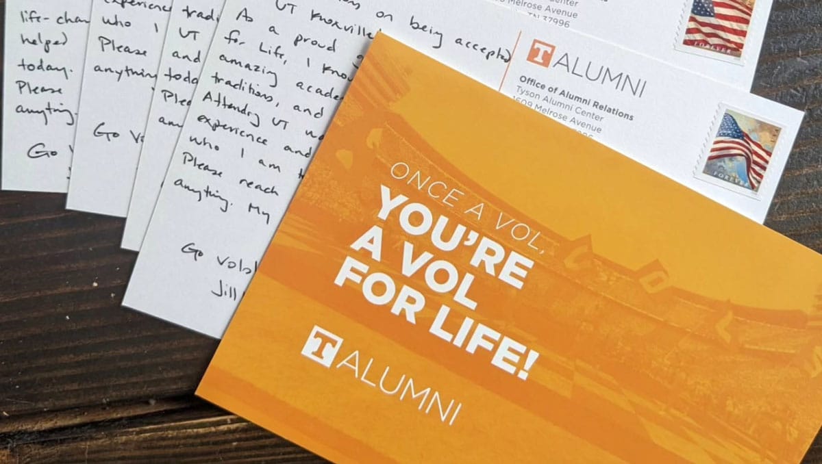 A stack of 5 postcards splayed on a table. The top postcard is orange and says You're a Vol For Life!