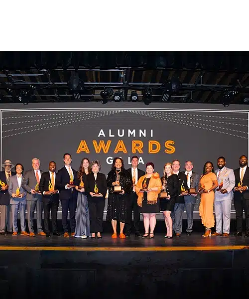 alumni-awards-winners-24 Sixteen Alumni Award winners stand together on a stage, holding their awards.