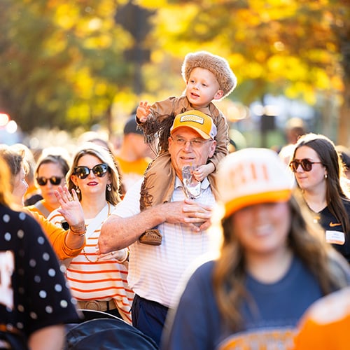 homecoming-parade-little-vol-walk-2 Families walk during the Little Vol Walk of the Homecoming parade. The focus is on a man with a young child dressed like Davy Crockett on his shoulders.