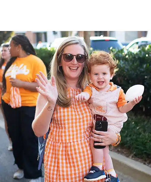 homecoming-parade-little-vol-walk A woman holding a child, and they both are waving at the camera. The child holds a white football toy.