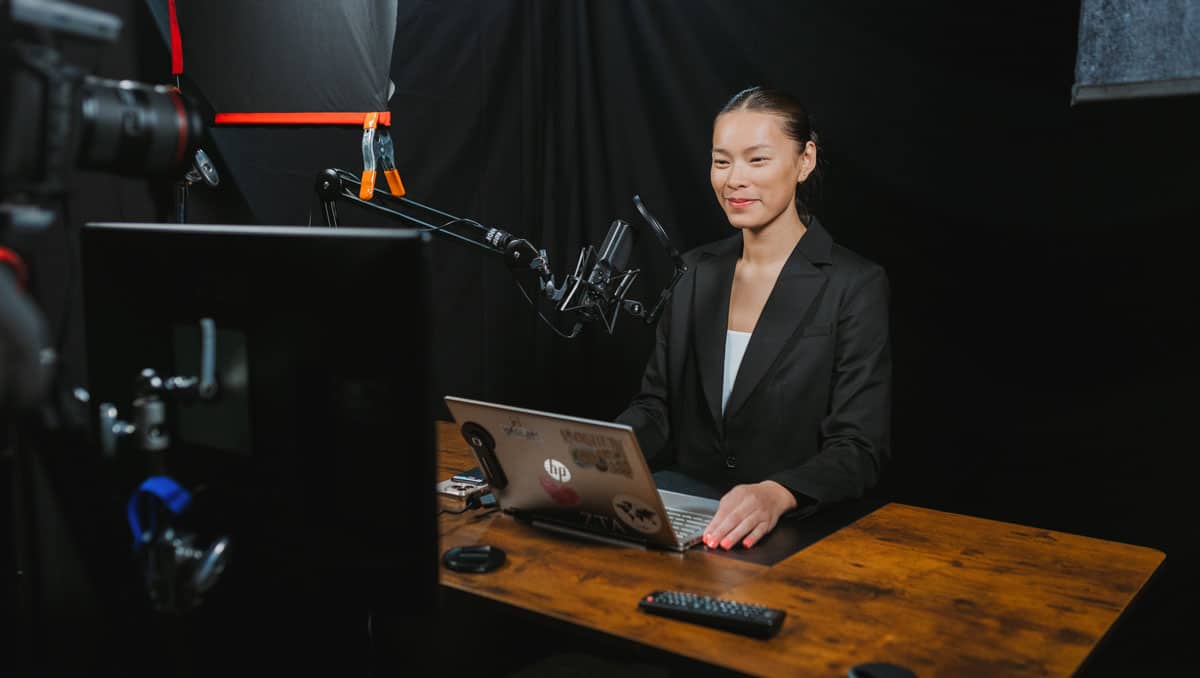 phelps-tatiana-recording-1200x678 Tatiana Phelps sits at a wooden table, a laptop, video camera, and microphone in front of her.