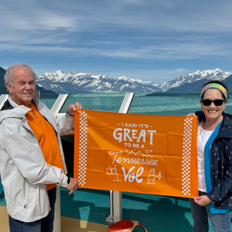 vantrease-jerry-and-jana-cruise-800x800 Jerry Vantrease and Jana Vantrease hold an orange It's Great to be a Tennessee Vol flag while on a cruise ship. In the background are snowcapped mountains.