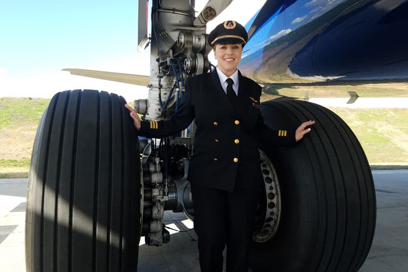 vieten-katrina-with-airplane-800x533 Katrina Vieten, wearing a pilot uniform, stands between the wheels of a commercial airplane.
