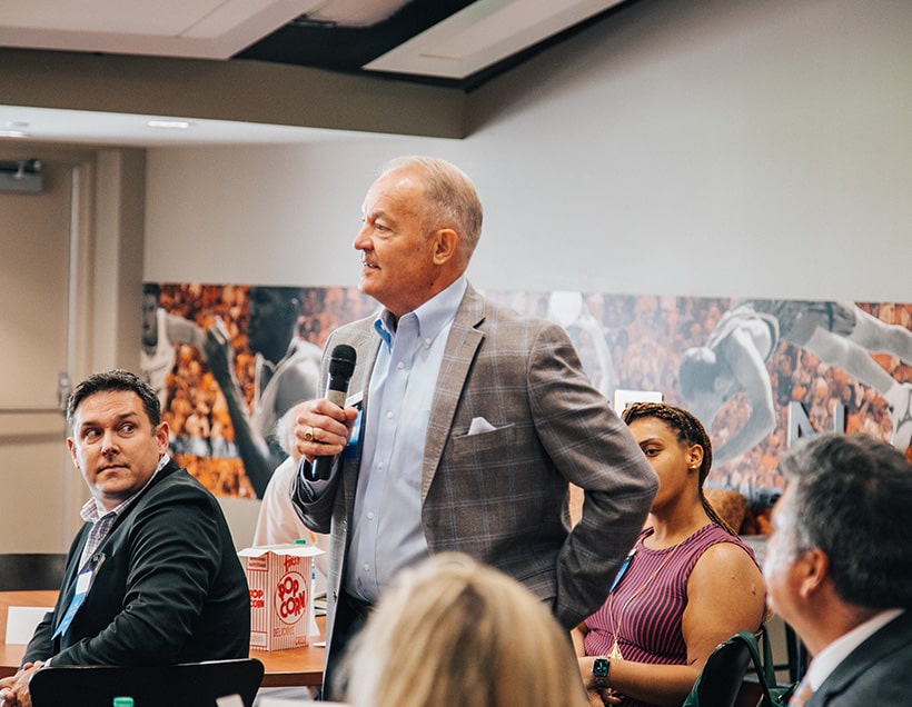 chancellors-assoc-opt-1 A man in a suit, surrounded by other people, stands and holds a microphone.