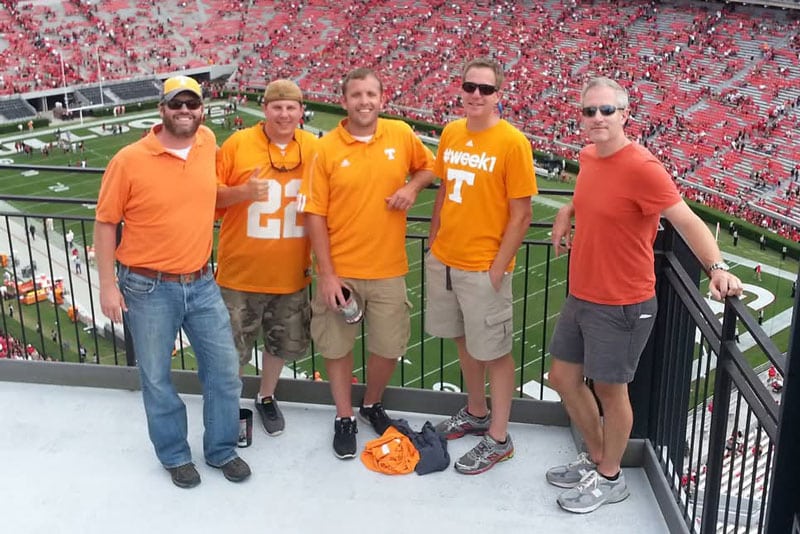 Five friends dressed in Tennessee orange pose for a photo with a football field behind them.